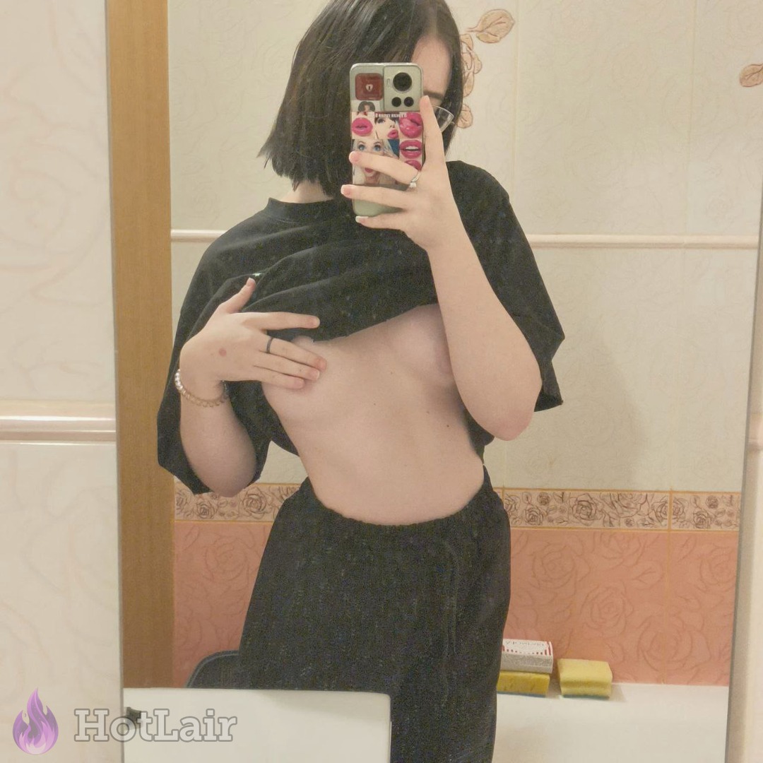 Selfie en espejo de una con cabello corto oscuro levantando su camiseta negra para mostrar sus pechos en el baño