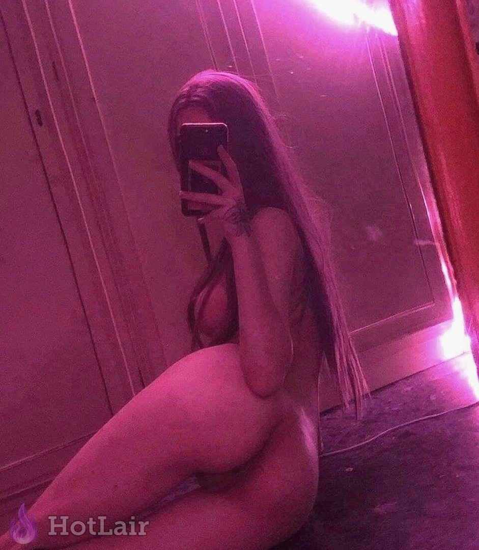 Mujer desnuda con largo cabello oscuro haciéndose selfie en espejo con luz rosa neón