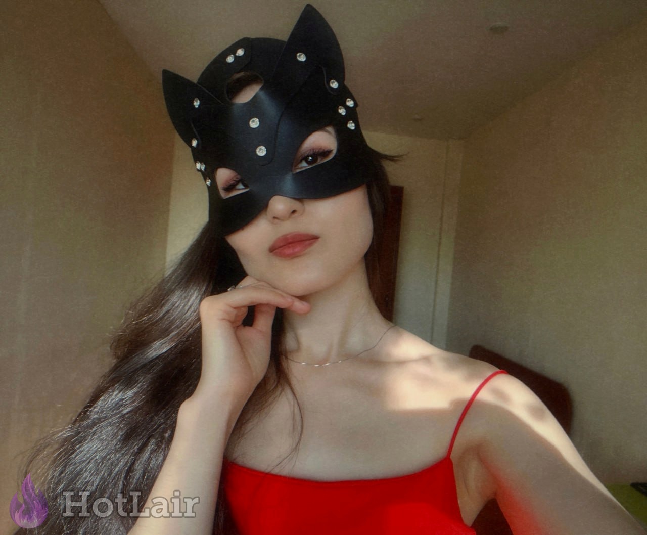 mujer asiática de cabello largo oscuro con máscara negra de gato con cristales y top rojo de tirantes finos, posa para selfie con mano en la barbilla