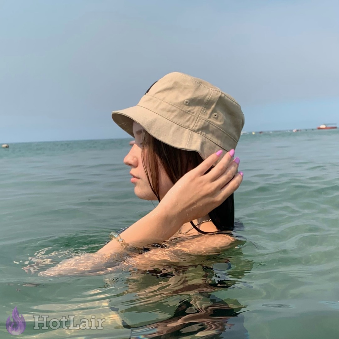 mujer con sombrero de pescador beige de pie en el mar con mano tocando el cabello