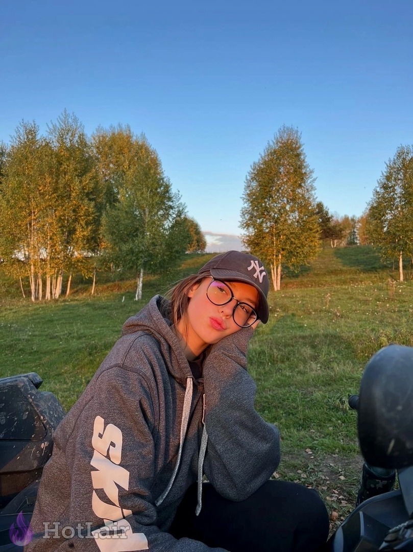 Jeune femme à lunettes et casquette posant dans un champ d’herbe au coucher du soleil en sweat gris