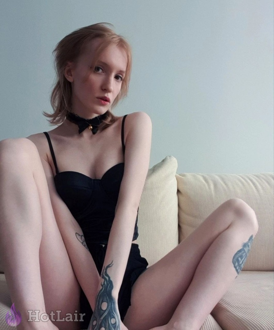 de cabello claro en corsé negro y choker con lazo sentada en sofá con rodillas arriba, tatuajes en brazos y piernas