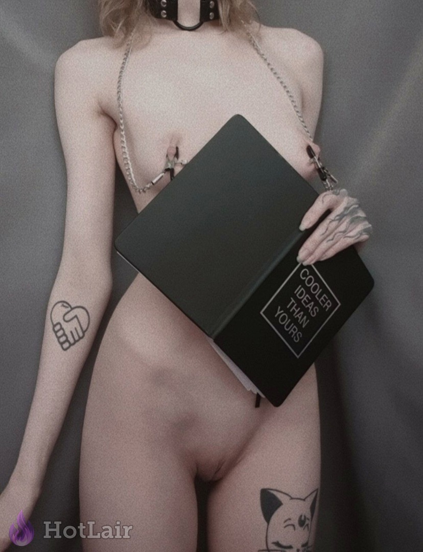 Desnuda rubia con collar BDSM, cadena y pinzas en pezones sosteniendo cuaderno negro, tatuajes visibles