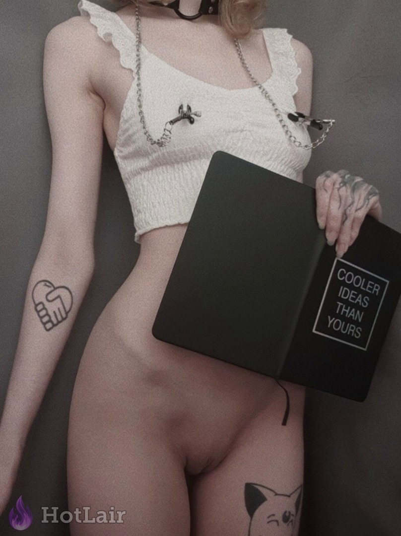 Mujer en top blanco con volantes y pinzas en pezones con cadena sosteniendo cuaderno negro “COOLER IDEAS THAN YOURS”, tatuajes visibles