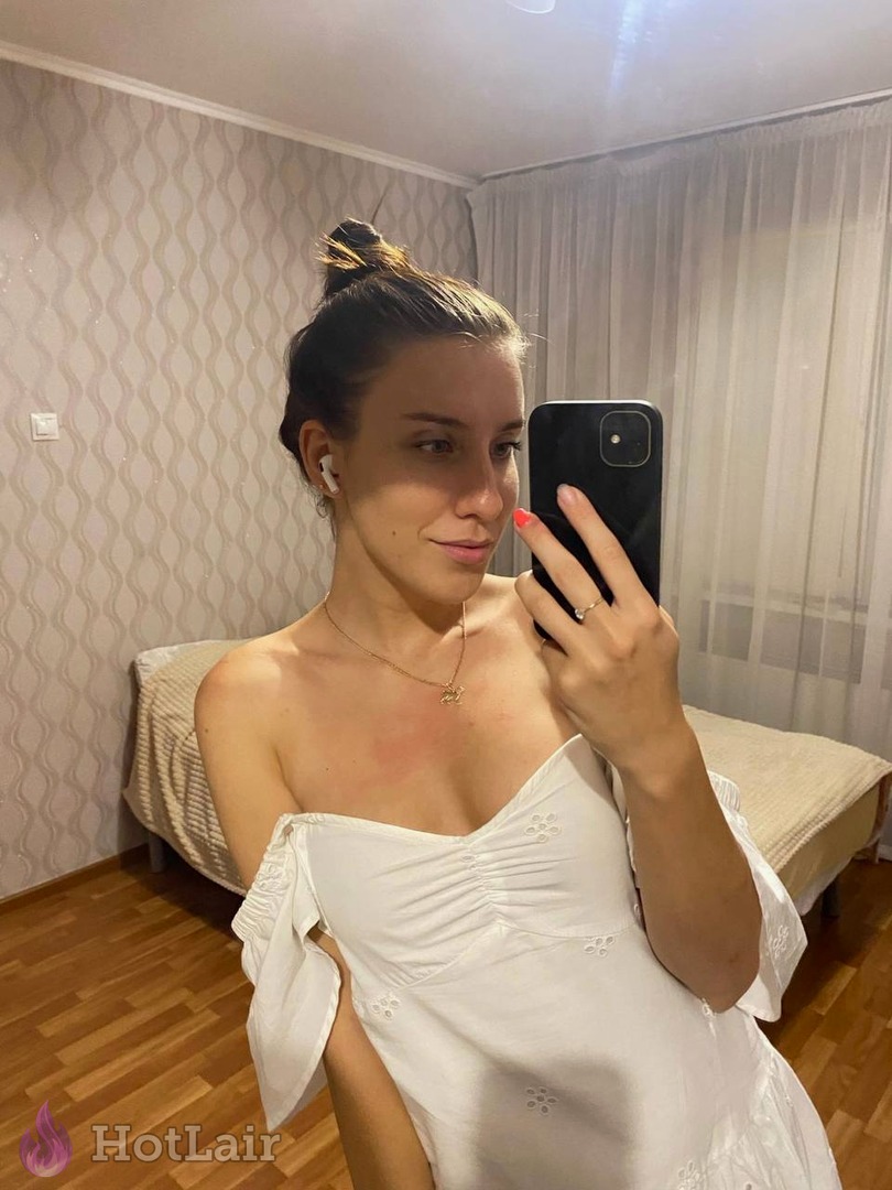 Jeune femme aux cheveux noirs en chignon prenant un selfie miroir en robe blanche décolletée tenant un smartphone noir dans une chambre