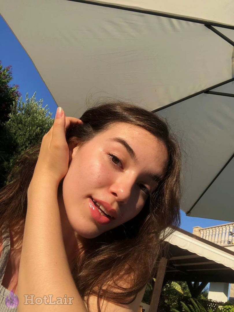 Jeune femme aux longs cheveux châtains souriant et touchant ses cheveux en selfie en extérieur sous parasol