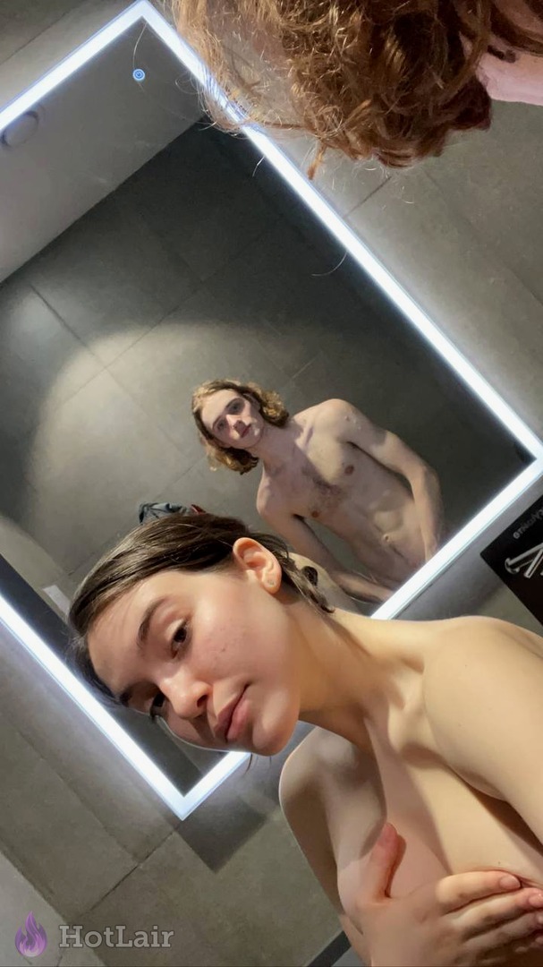 Femme prenant un selfie miroir dans salle de bain moderne avec éclairage LED, couvrant son sein, reflet d’une autre personne visible