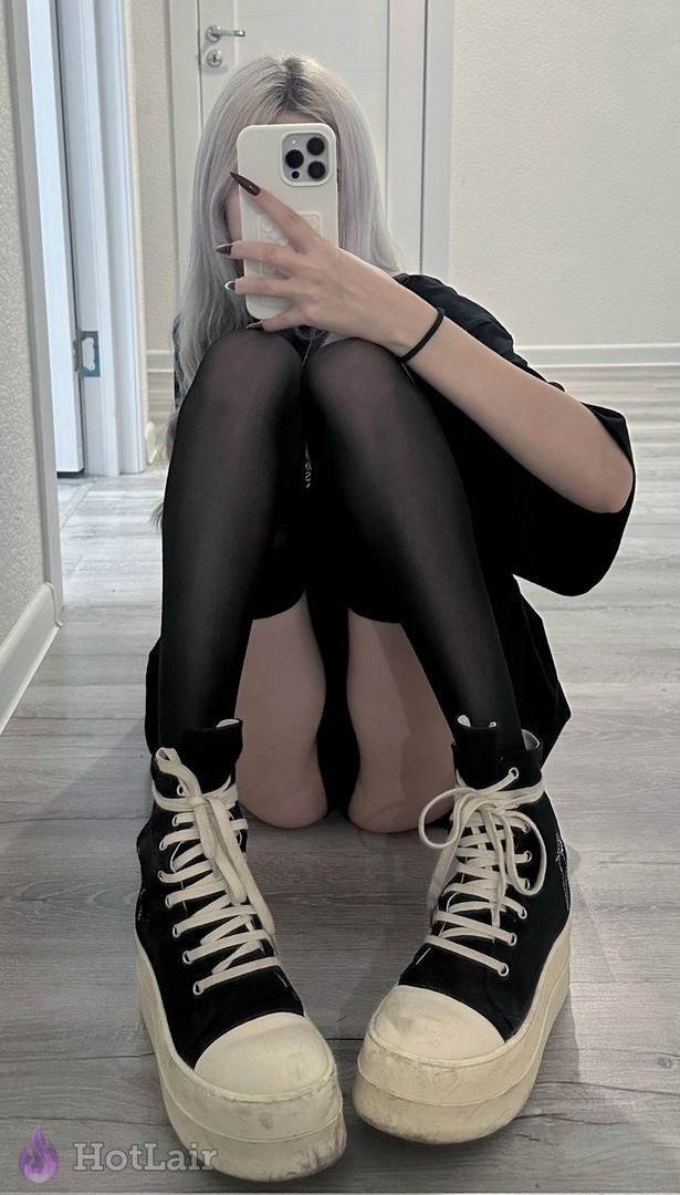 de cabello platino sentada en el suelo con medias negras y camiseta oversize negra, zapatillas plataforma grandes, haciéndose un selfie con el teléfono cubriendo su rostro