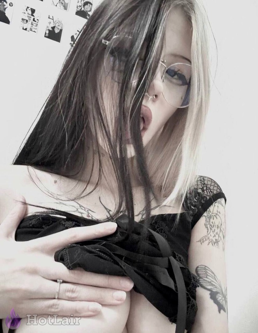 Chica alternativa tatuada con cabello bicolor negro y rubio, gafas y piercing en la nariz bajando su top de encaje negro para mostrar los pechos, selfie close-up con lengua afuera