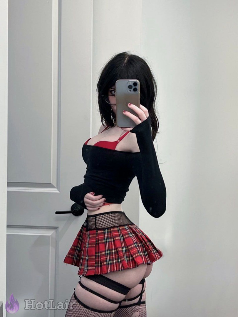 Chica de cabello negro con top negro off-shoulder y falda plisada roja a cuadros con liguero haciéndose selfie en espejo