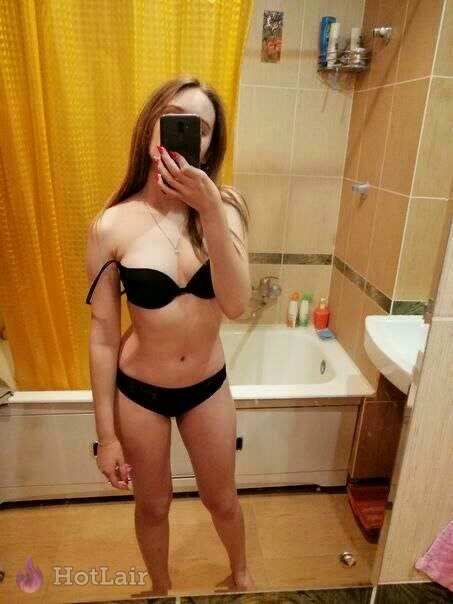 con largo cabello castaño haciéndose selfie en espejo en el baño con sujetador y bragas negras, sosteniendo smartphone con uñas rojas