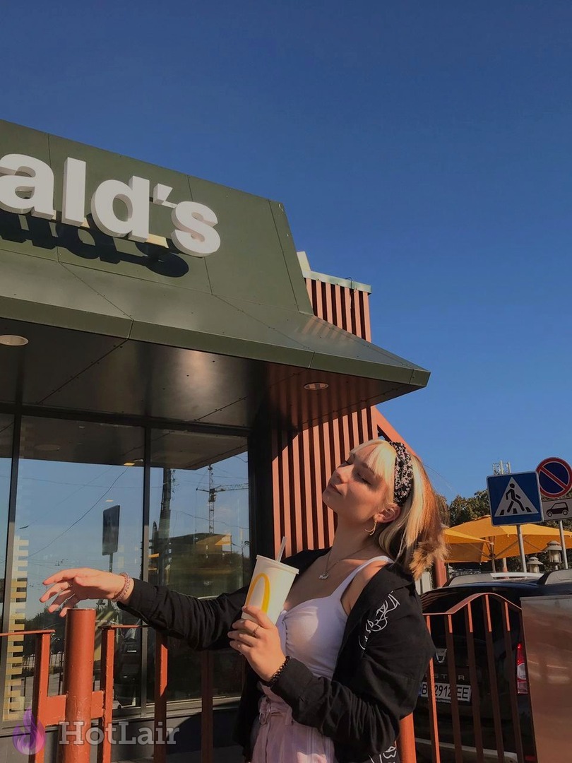 Blonde posant devant un McDonald’s sous un ciel bleu clair, tenant un gobelet