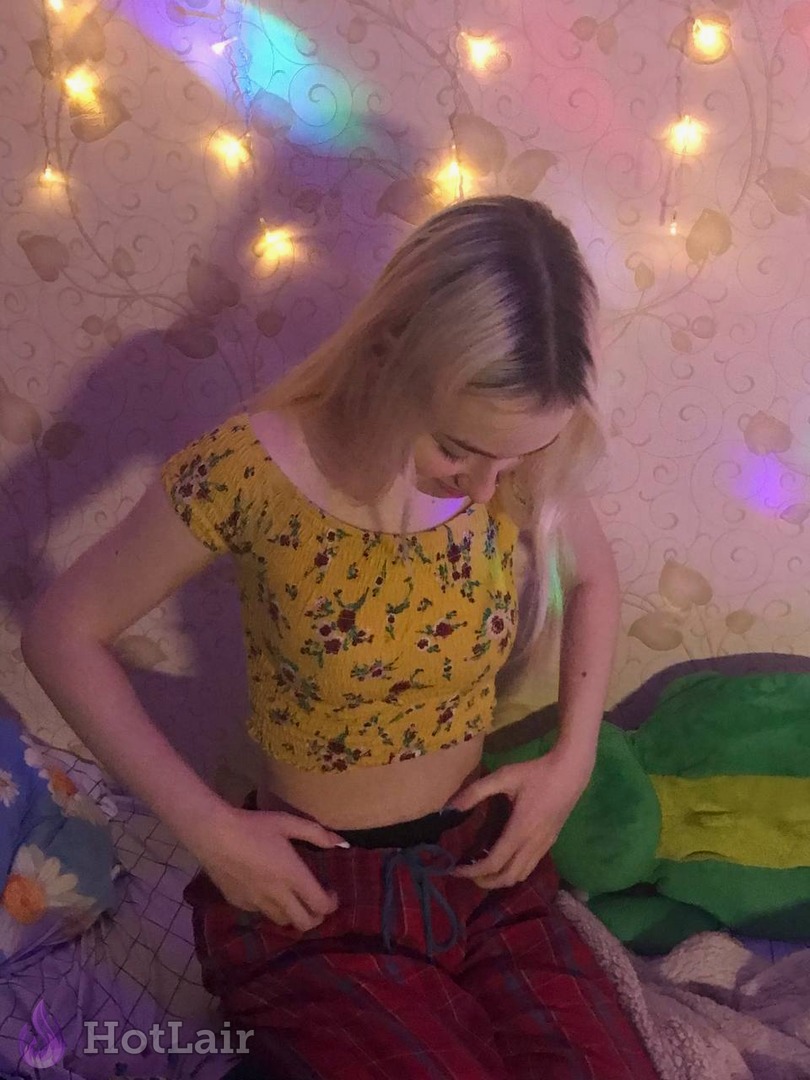 Blonde en crop top jaune fleuri baissant un pantalon à carreaux rouge dans une pièce cosy