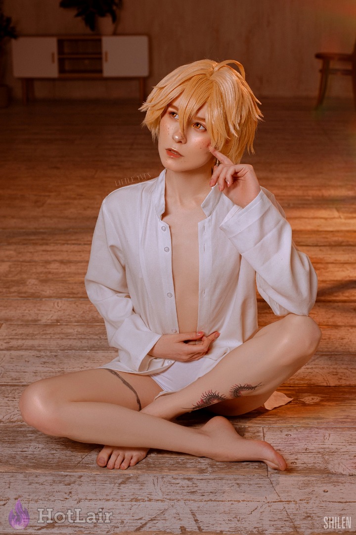 Blonde androgyne en chemise blanche déboutonnée assise pieds nus sur parquet, doigt sur la joue