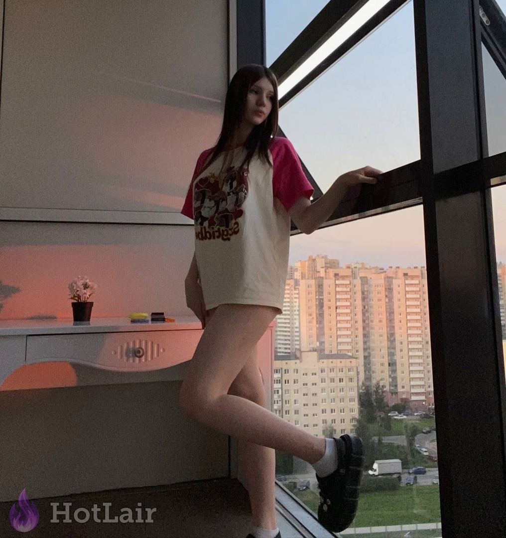 con largo cabello castaño en camiseta oversize posando junto a ventana grande al atardecer