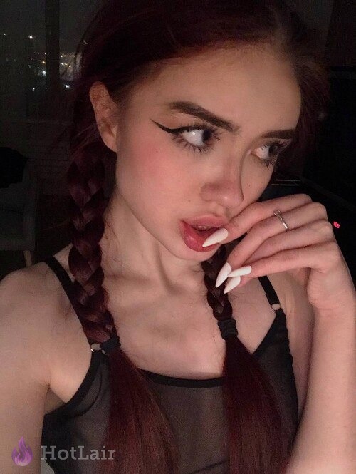 Mujer con trenzas y cabello rojo en top negro transparente posando con dedo cerca de la boca