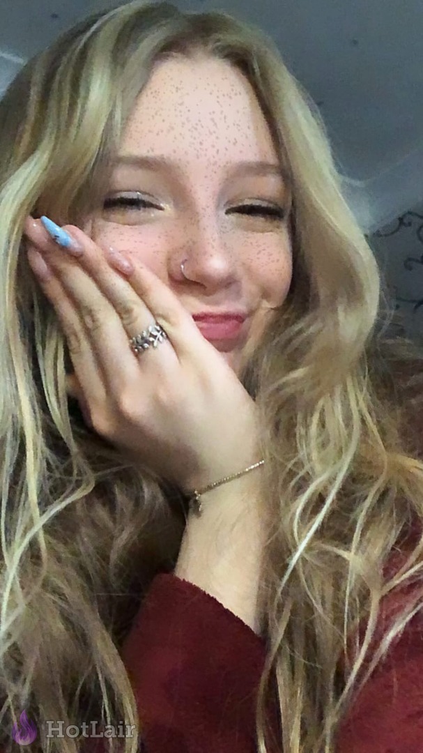 Rubia sonriente con pecas y piercing en la nariz apoyando la mano en la mejilla