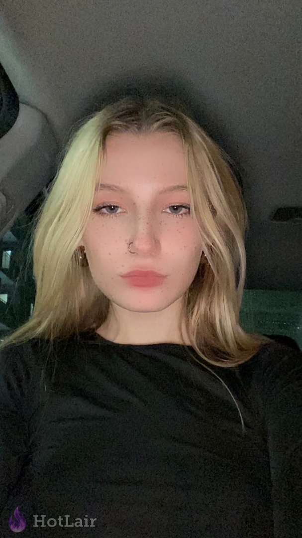 Selfie cercano de rubia con pecas y piercing en la nariz dentro de un coche, con top negro