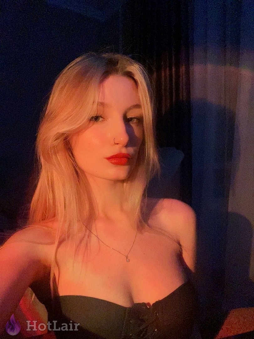 Primer plano seductor de rubia en top corsé negro con labial rojo bajo luz rosa cálida, escote profundo