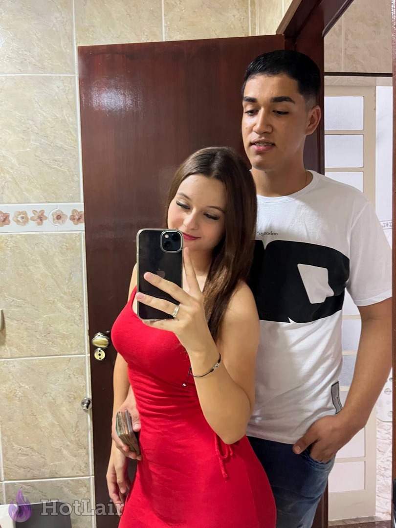 Pareja haciéndose un selfie en el espejo: morena con vestido rojo sosteniendo un smartphone negro y un hombre detrás