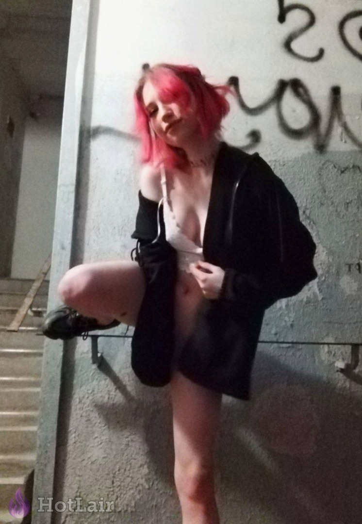 Mujer de cabello rosa de pie contra pared de grafiti con chaqueta negra abierta y lencería blanca